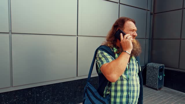 Long-haired bearded senior man stands near a building and calls someone