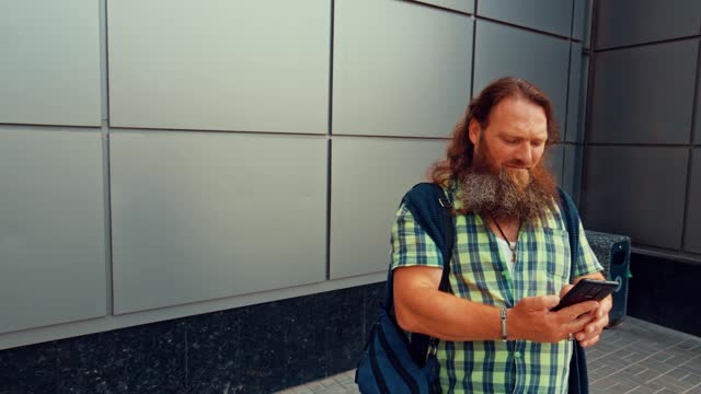 Bearded long-haired man is trying to call someone while standing near a building