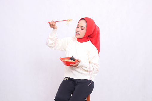 candid asian woman wearing hijab looking down holding noodles with chopsticks from the side and bowl containing ramen (chinese food). Beautiful Muslim women use culinary, food and health content