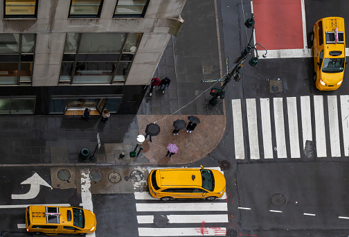 Aerial view photography of New York City.  This is on a very wet and dark spring day with wet roads and three yellow cabs at a cross roads.   There are people carrying umbrellas on the side walk.