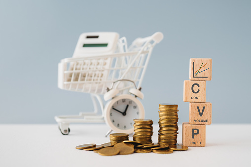 CVP text ,cost, volume, profit , and graph on wooden cube blocks, with stack of coins , blurred clock ,calculator, shopping cart. Business concept