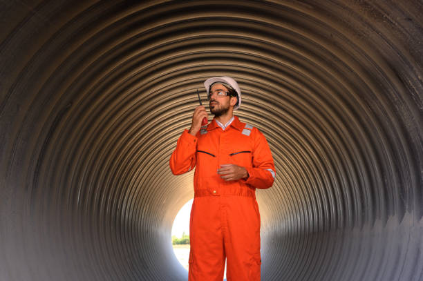 An engineer in a reflective orange jumpsuit and hard hat stands inside a corrugated metal tunnel using a walkie-talkie.