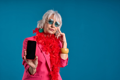 tech-savvy pensative senior lady in trendy pink with smartphone and blue background