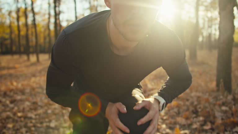 Close up a man in a black sports uniform ties the laces on his sneakers before starting his race through the autumn sunny forest in the morning. Confident man with curly hair starts his run in the morning in the forest