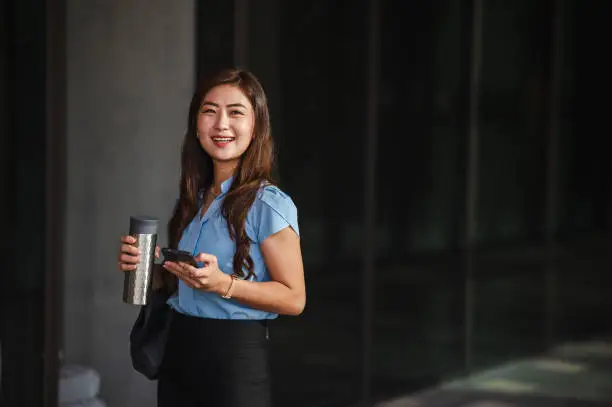 An Asian businesswoman is using her smartphone while holding a water bottle in the city. She's conducting business on the move. Looking at camera.