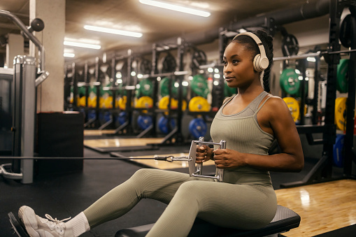 A powerful black sportswoman with headphones practicing backs on seated cable machine at gym.