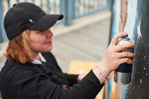 Female artist in black cap is painting picture with paint spray can spraying it onto canvas at outdoor street exhibition, side view of female art maker