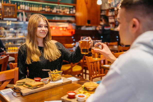 young couple cheering with beer while eating burgers and sandwiches in a pub - toast portion club sandwich cafe photos et images de collection