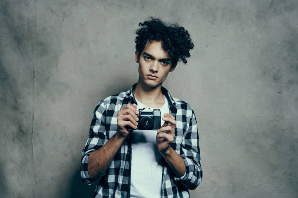 Cool stylish man professional photographer with glasses in a beard with a tattoo in a fashion knitted hat and sweater holds a modern mirrorless photo camera in the studio