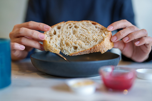 In the cropped image, an Asian woman is seen enjoying her breakfast in a cafe during the morning. She has sourdough bread on her plate, along with butter and strawberry jam.