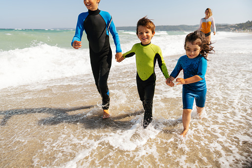 Photo of a single mother with three children in wetsuits, walking down the beach and playing with the ocean waves
