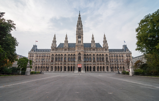 Vienna, Austria - October 10, 2013: symmetric view of neo-gothic Rathaus building and empty square in front of it, green trees on the sides. Cloudy day.