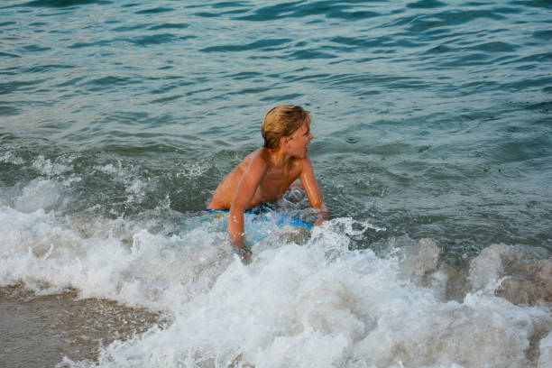 A young boy stands by the water's edge at the beach, holding a bright surfboard under the sun. People relax nearby as waves gently lap at the shore, capturing summer vibes.