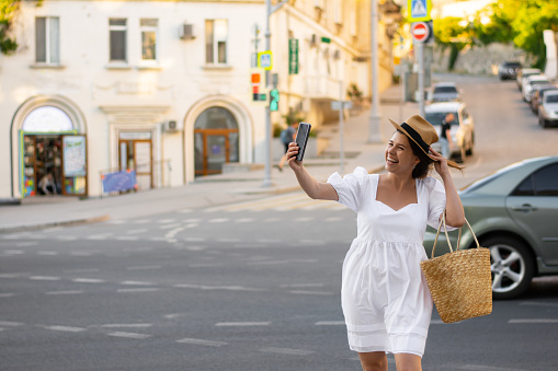 Charming woman smiling with white teeth tourist taking a selfie on her phone in a European city. Architecture, tourist travel, lifestyle, trendy clothes, woman in white dress, straw hat, straw bag