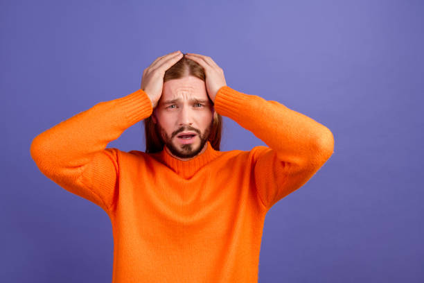 portrait d’un jeune homme barbu drôle contrarié dans les mains de chandail orange touchent la tête choquée et perplexe isolé sur fond de couleur violette - long blond hair photos et images de collection