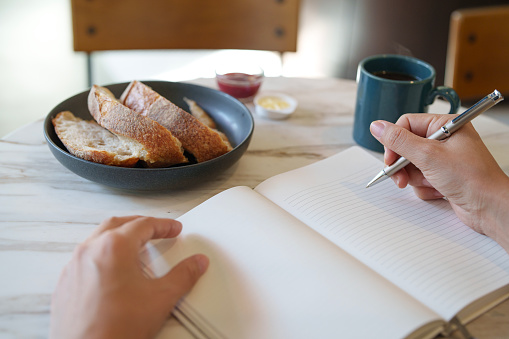 Close-up of an Asian woman at a table writing on a notepad while enjoying breakfast with coffee and sourdough bread in the morning, creating a balanced and productive workspace