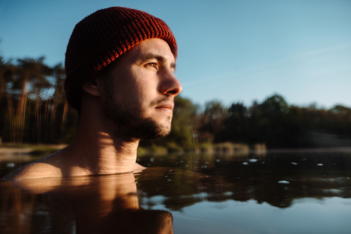 Young man in hat bathing in the ice water alone at the morning. Boy or man bathing and swimming in the cold water of a lake or river. Cold therapy and ice swim concept