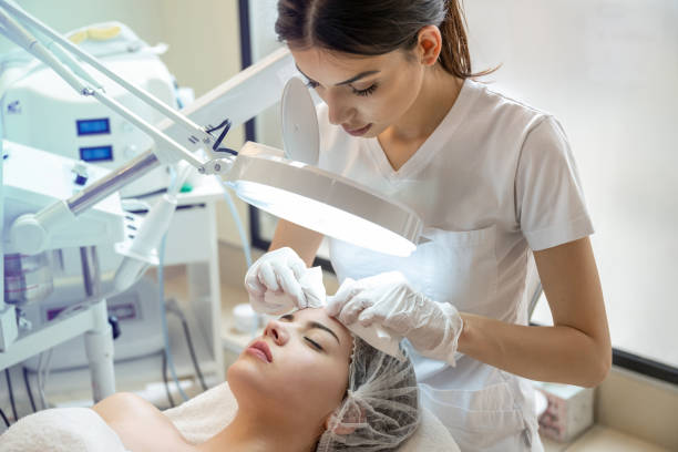 istock Beautician squeezing acne and pimples from young woman's nose using lamp and magnifying glass in beauty spa clinic. 2083623273