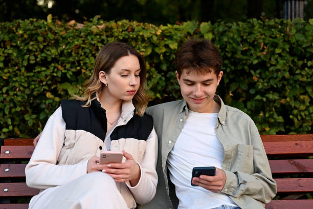 istock A jealous woman peeks at her boyfriend's phone while relaxing on a park bench 2083637215
