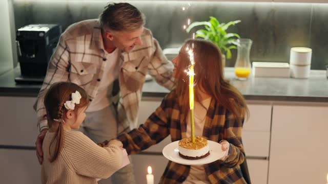 A little brown-haired girl and her middle-aged dad with gray hair congratulate their mom and brunette wife in a checkered shirt and bring her a cake with a huge candle during dinner at the dining table in the evening in a modern apartment