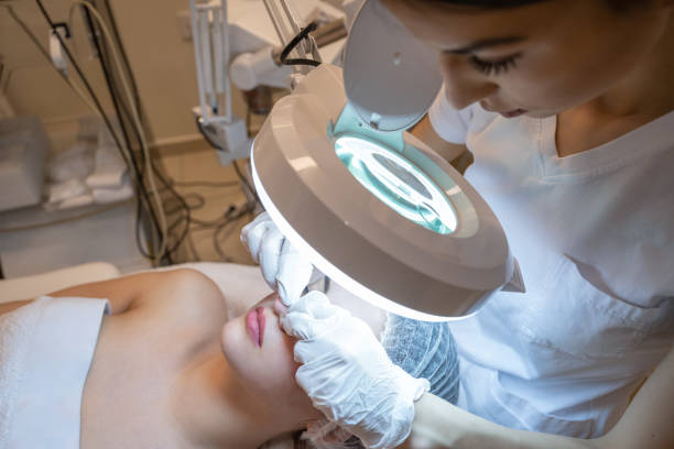 istock Beautician squeezing acne and pimples from young woman's nose using lamp and magnifying glass in beauty spa clinic. 2083668008