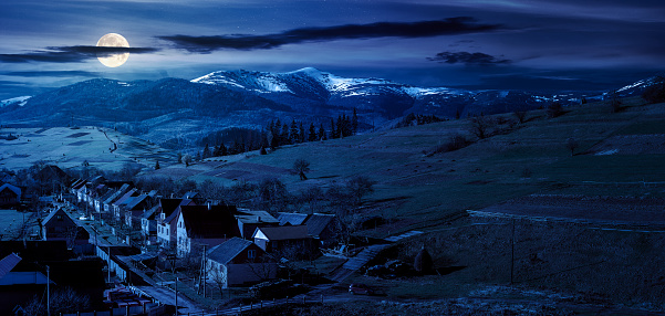 alpine countryside ant night. village in the valley and distant mountain range with snow capped tops in full moon light