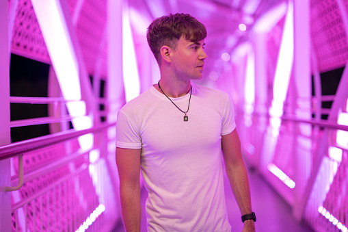 Side view portrait of attractive handsome young man wearing white shirt posing at pedestrian overpass with pink lights