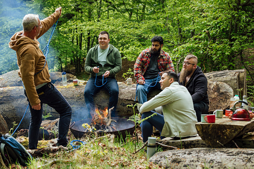 Group of men sitting around a fire pit in a woodland area in Northumberland, North East England. A man is teaching the rest how to tie a knot with rope.

Videos available for this scenario.