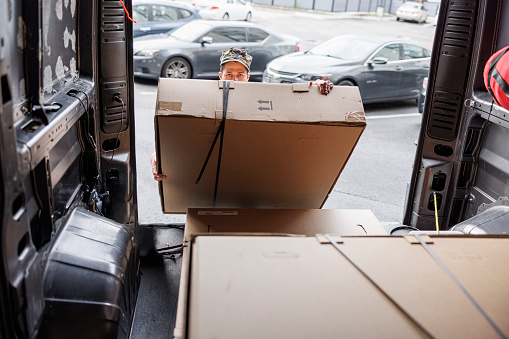 A young Hispanic Navy soldier efficiently unloads the cargo van, retrieving a heavy box. Looking from the van, we can see only his eyes staring at camera and a cap above the box