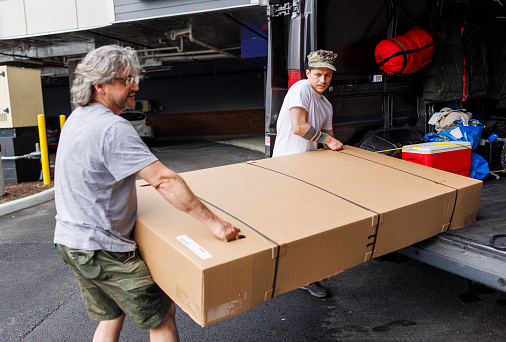 Young Latino Navy officer and mature Caucasian gray-haired man in eyeglasses, carrying heavy wrapped box from cargo van