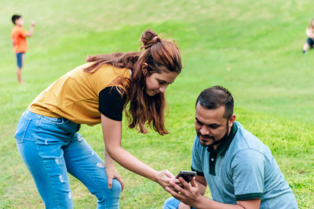 młoda latynoska kobieta pokazuje swój telefon swojemu chłopakowi, siedzącemu w parku. - contreras zdjęcia i obrazy z banku zdjęć