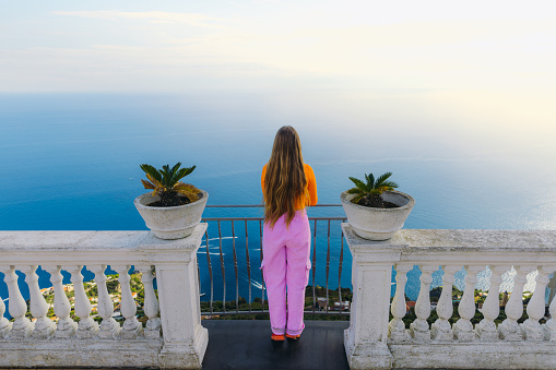 Rear view of smiling female in orange shirt walking at the viewpoint of Amalfi Coast of Italy admiring view of blue sea