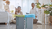 Happy Asian senior couple play garbage sorting game, throw plastic bottle to litter recycle bin labeled recyclable, general waste. Eco-friendly home, waste separation management, sustainable lifestyle