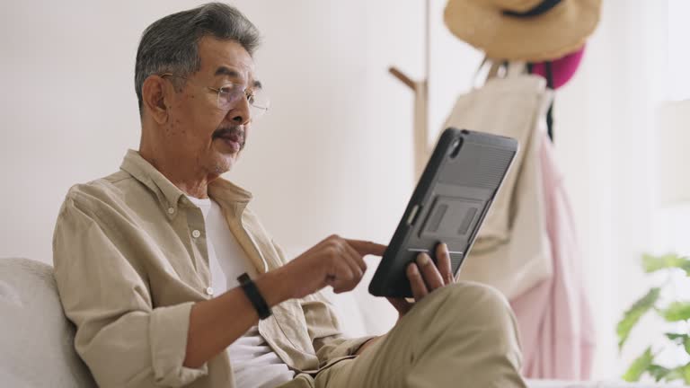 Senior male using digital tablet for serving the internet on sofa at home. Connecting at home after retirement.