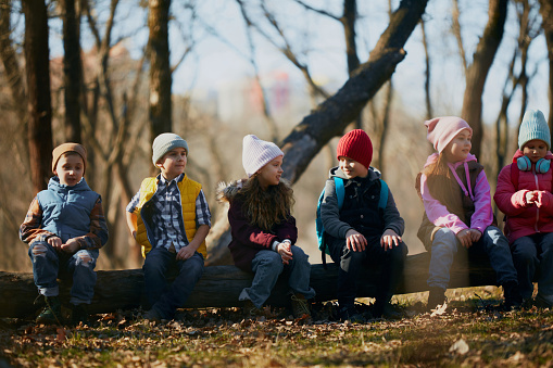 Row of kids, classmates perched on a log, clad in colorful winter wear, enjoying lesson in forest. Concept of outdoor activities for children's development, school, childhood, fashion and style. Ad