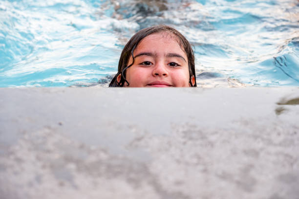 Young girl in pink swimsuit swimming in clear blue pool water, gazing at the camera with a serious expression. Fun, summertime, and outdoor activity for kids.