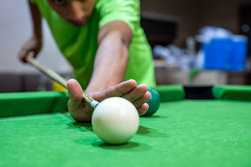 Young adult bearder male playing billiards with his friends focusing on white ball to hit it with muscular hand