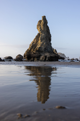A large rock sits on a beach with the ocean in the background. The rock is a reflection of itself in the water