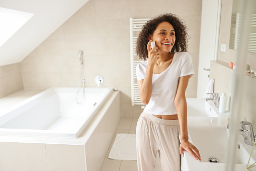 A woman in the buildings bathroom, brushing teeth with one hand while talking on a cell phone. Window overlooks the street, wood flooring under her feet