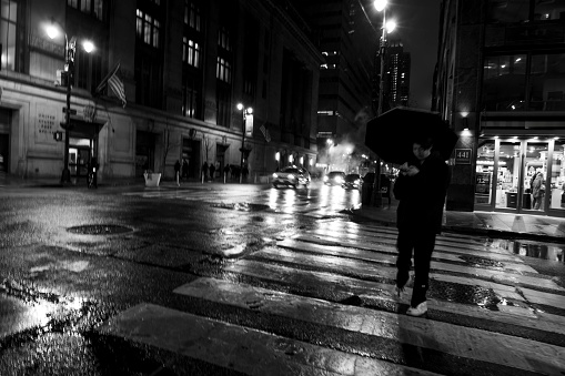 Manhattan, New York, USA - March 2024. Street photography at night in Manhattan.  In the foreground is a man crossing a zebra crossing holding an umbrella.