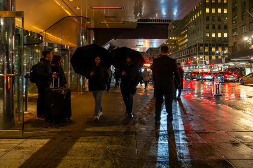 Manhattan, New York, USA - March 2024. Street photography at night in Manhattan.  In the foreground are people outside a hotel, some with umbrellas.