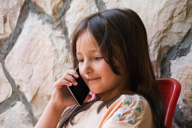 Cheerful little girl using smart phone while sitting alone in garden yard . Little girl having online chat with her parents