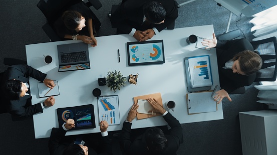 Top down aerial view of smart manager pointing at financial graph on whiteboard while diverse business team explain and plan marketing idea at meeting table with stock market statistic. Directorate.