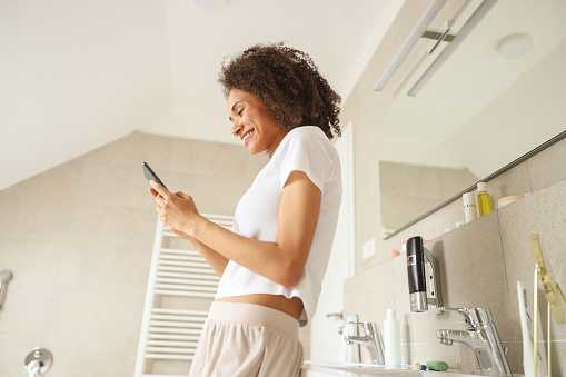 A woman, standing on the hardwood floor, gazes at her phone in the bathroom. Her elbow rests on the window ledge as she scrolls, while the ceiling looms above her head