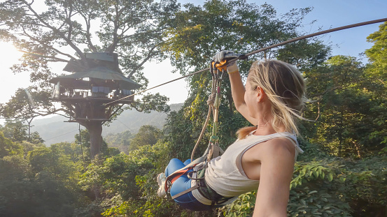In the heart of the lush jungle canopy in Laos, a young woman embarks on an exhilarating adventure, zip lining through the verdant foliage. Suspended from a sturdy cable, she gracefully glides through the emerald treetops, her laughter echoing amidst the rustling leaves and the distant calls of exotic birds. With each thrilling descent, she embraces the rush of adrenaline, her spirit soaring in harmony with the untouched wilderness surrounding her. Against the backdrop of towering trees and cascading waterfalls, she embraces the freedom and serenity of this immersive jungle experience, a fleeting moment of pure joy captured amidst the untamed beauty of nature.