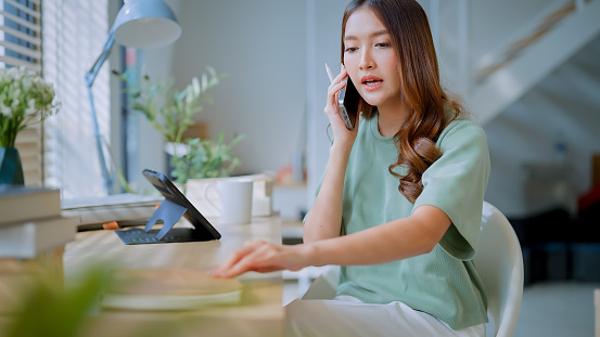Smiling freelancer Asian female adult woman sitting on chair working with laptop tablet on in living room window morning casual lifestyle ,Working From Home in Cozy Sunny Atmosphere