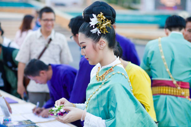 Profile portrait of elegant traditionally dressed thai woman. She has  flower decor  in hair. Woman is wearing traditional elegant clothes. Top is turquoise colored.  In background are more people in traditional fashion including blue farmer style. Scene is at a local festival with market near to King Naresuan monument in Lampang. People in background are filling out some survey papers.