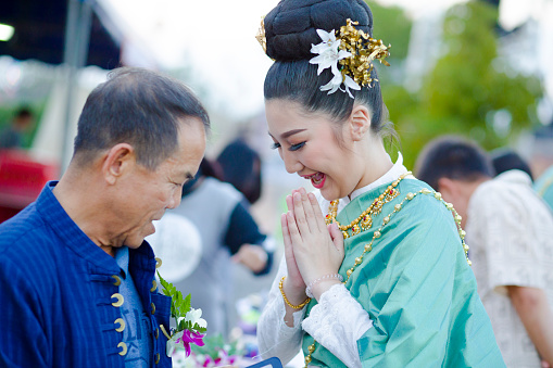 Traditionally dressed thai woman is thanking mature man with wai gesture. People in Lampang. There are a mature man in in blue fashion and a young woman with flower in hair in turquoise colored clothes,. Woman has placed some small flower decor at chest and jacket of man and is adressing him respectfully..   Scene is at a local festival with market near to King Naresuan monument in Lampang.