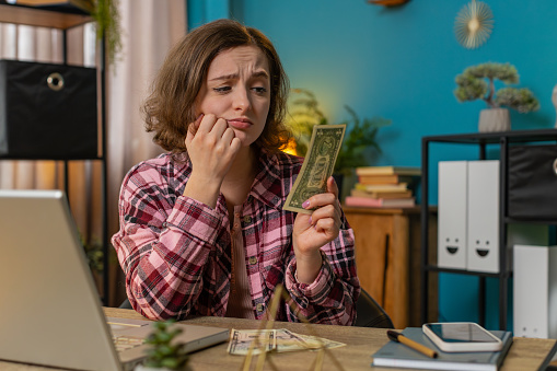 Caucasian young woman freelancer counting cash insufficient amount of money. Financial crisis. Bankruptcy. Poverty and destitution. Female girl businesswoman with laptop sitting at home office table.