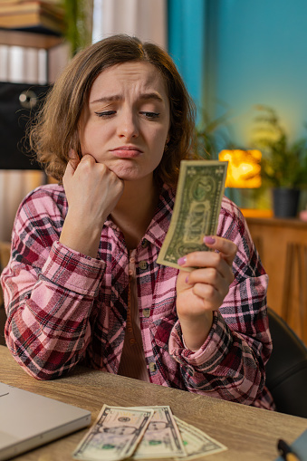 Young woman freelancer counting cash insufficient amount of money. Financial crisis. Bankruptcy. Poverty and destitution. Female girl businesswoman with laptop sitting at home office table. Vertical